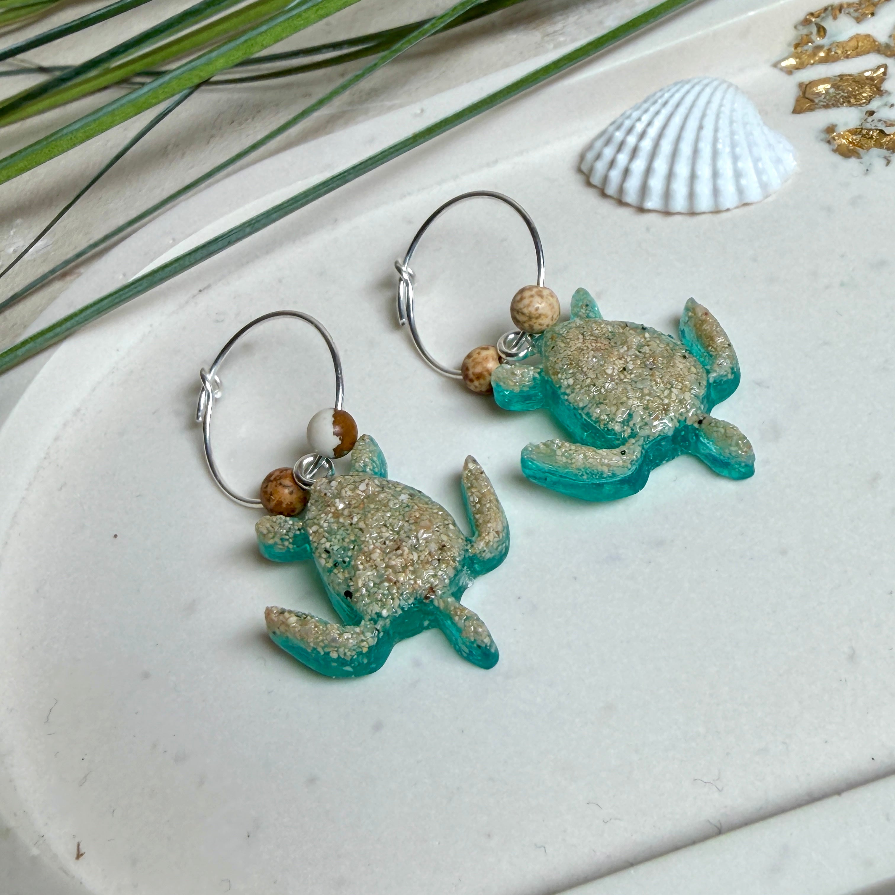 Türkisfarbene Schildkröten-Creolen an feinen 16-mm-Ringen aus 925 Silber, jeweils mit echtem Strandsand im Harz eingebettet und mit kleinen Jaspis-Edelsteinperlen verziert; handgefertigtes Unikat mit sommerlich-maritimem Look, fotografiert als Freisteller, Nahaufnahme oder auf dekorativem Untergrund.