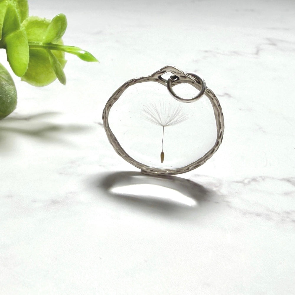 „Handgefertigter Pusteblumen Anhänger mit echter Löwenzahn Pusteblume in klarem Resin, runder silberfarbener Rahmen, filigraner Natur Schmuck auf hellem Marmor Hintergrund.“