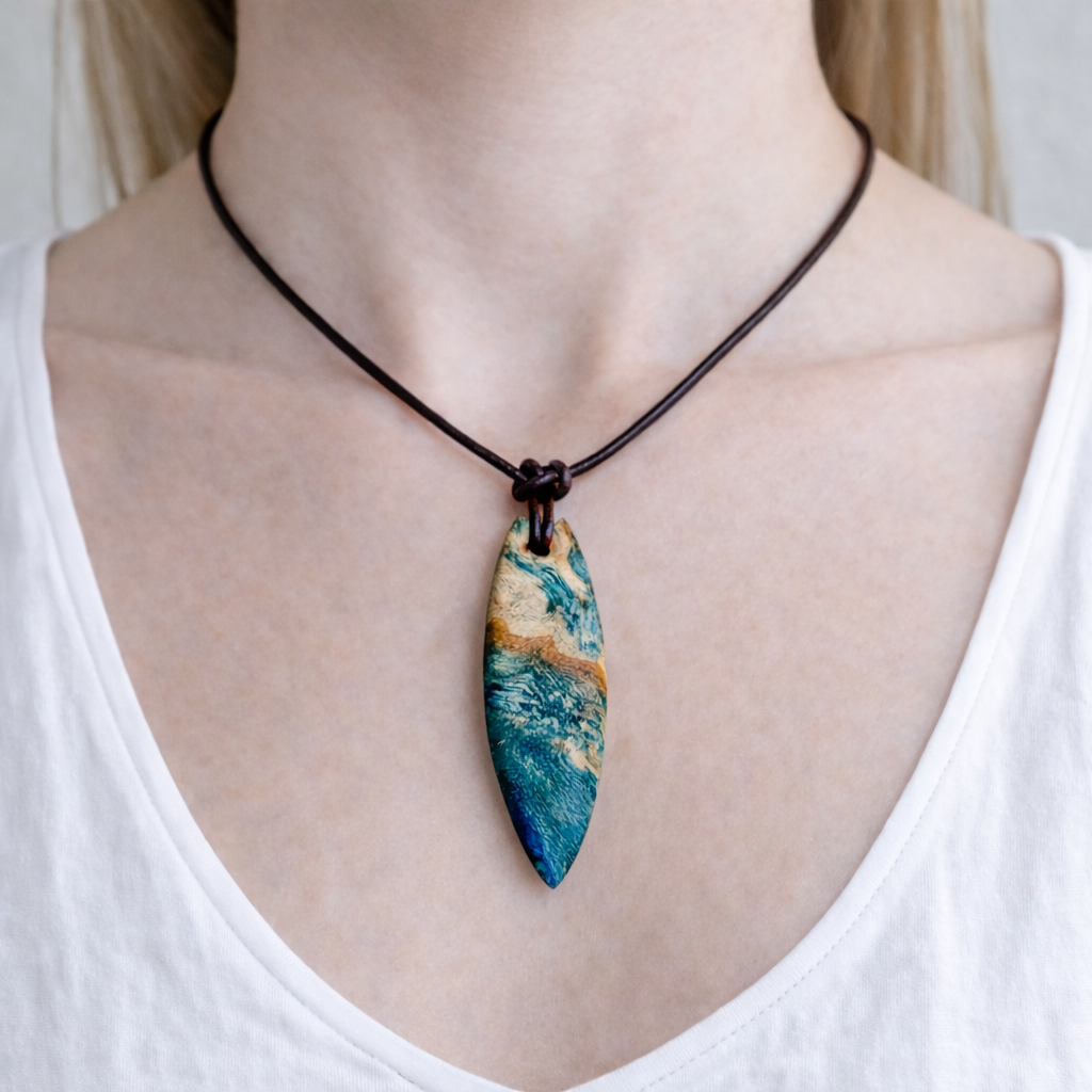Unikat Anhänger aus stabilisiertem Holz mit türkisblauem Harzverlauf, schlanke Form, poliert. Verstellbares braunes Lederband 22–46 cm mit Schiebeknoten. Anhänger ca. 6,6 × 2 cm. Tragefoto am Hals und Produktfotos mit maritimer Deko