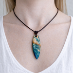 Unikat Anhänger aus stabilisiertem Holz mit türkisblauem Harzverlauf, schlanke Form, poliert. Verstellbares braunes Lederband 22–46 cm mit Schiebeknoten. Anhänger ca. 6,6 × 2 cm. Tragefoto am Hals und Produktfotos mit maritimer Deko