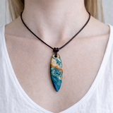 Unikat Anhänger aus stabilisiertem Holz mit türkisblauem Harzverlauf, schlanke Form, poliert. Verstellbares braunes Lederband 22–46 cm mit Schiebeknoten. Anhänger ca. 6,6 × 2 cm. Tragefoto am Hals und Produktfotos mit maritimer Deko