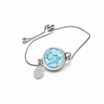 Verstellbares Edelstahlarmband mit Zirkoniasteinchen und Schiebeverschluss und rundem Anhänger (ca. 2,5 × 1,7 cm) mit echten Larimar-Edelstein-Splittern in Harz. Zusätzlich ein Charm „Blume des Lebens“ aus 925 Silber. Handgefertigtes Unikat in Türkis- und Meeresblautönen – Schmuckstück in Nahaufnahme bzw. als Tragebild am Handgelenk.