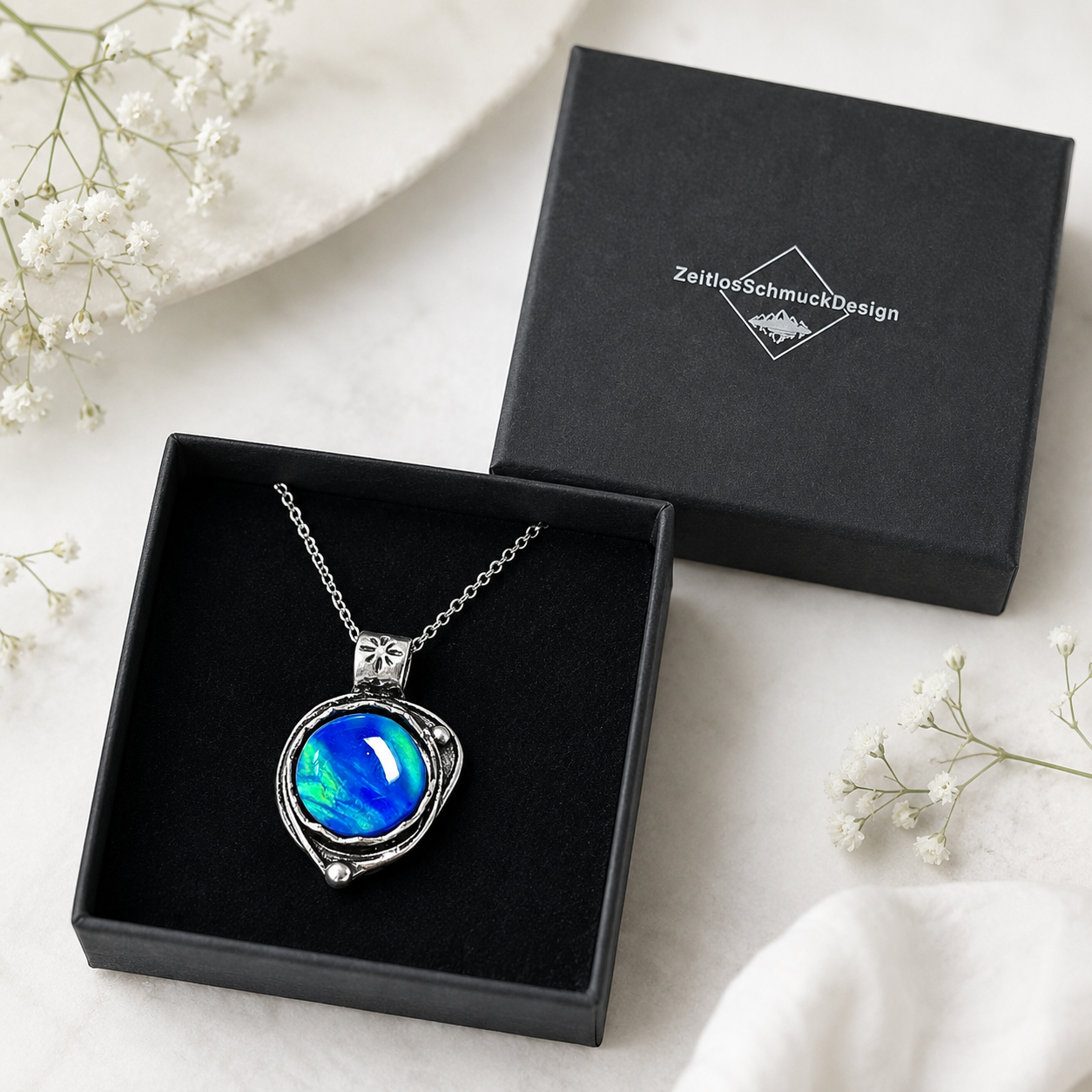 Labor-Opal-Anhänger „Galactic Glow“ in Antik-Silber-Optik mit blau-grün schimmerndem Cabochon liegt in einer geöffneten schwarzen Schmuckbox von ZeitlosSchmuckDesign.